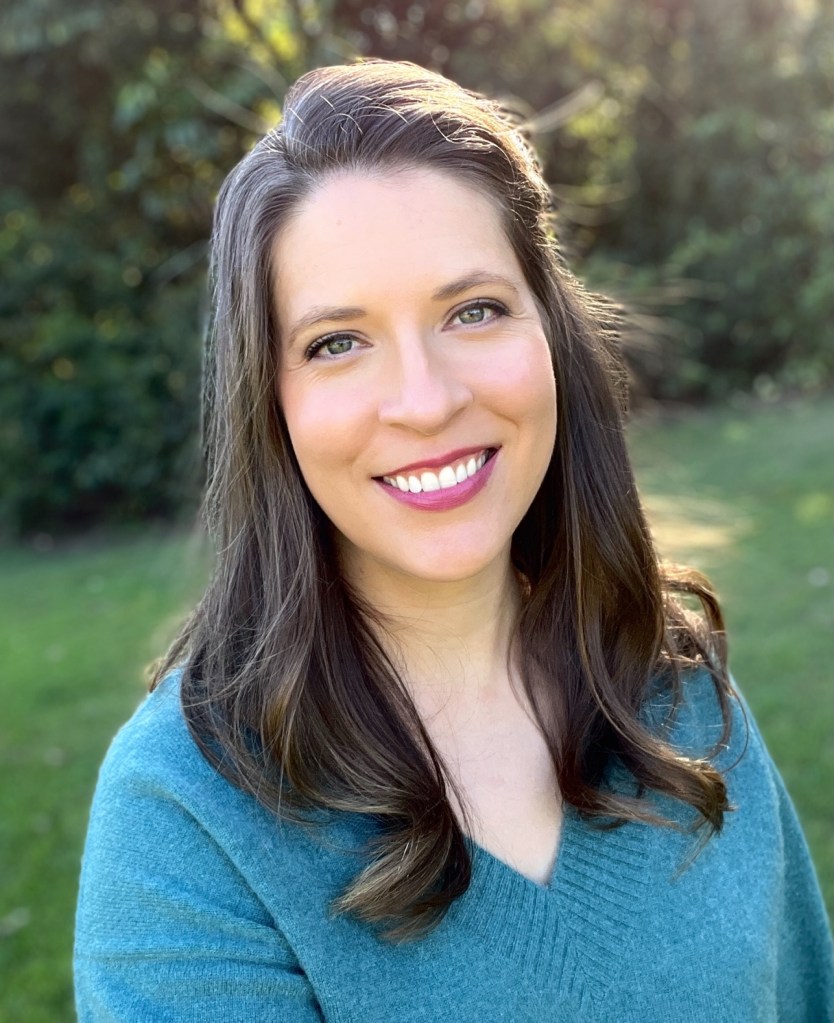 A photo of a woman in a teal jumper smiling at the camera. She is standing outside.
